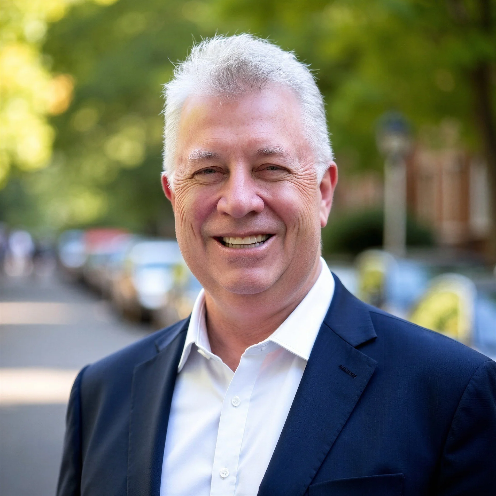 Close-up of a smiling man with gray hair, wearing a dark suit and white shirt, outdoors with blurred green trees in the background.