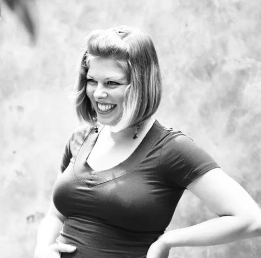 Black-and-white photo of a woman with short hair, smiling and looking to the side, wearing a fitted T-shirt, standing against a textured background.