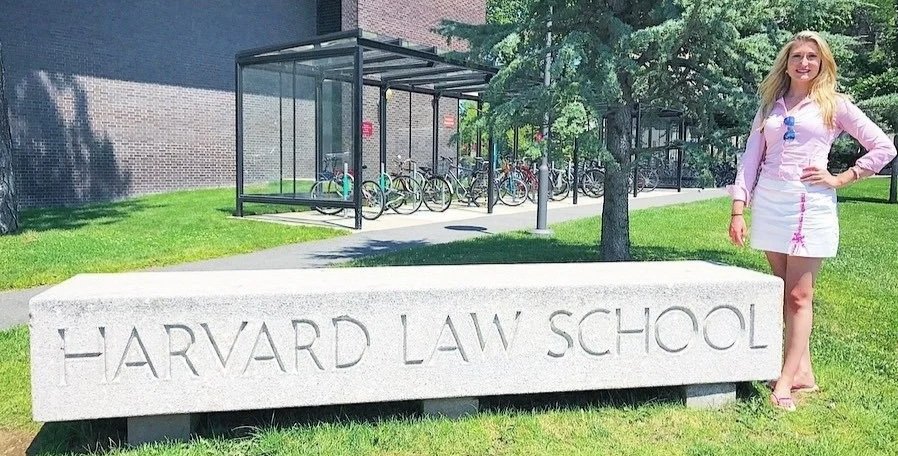 Caroline Shinkle, wearing a light pink top, white skirt, and sunglasses, standing next to a large stone sign that reads "Harvard Law School" outside on a sunny day.