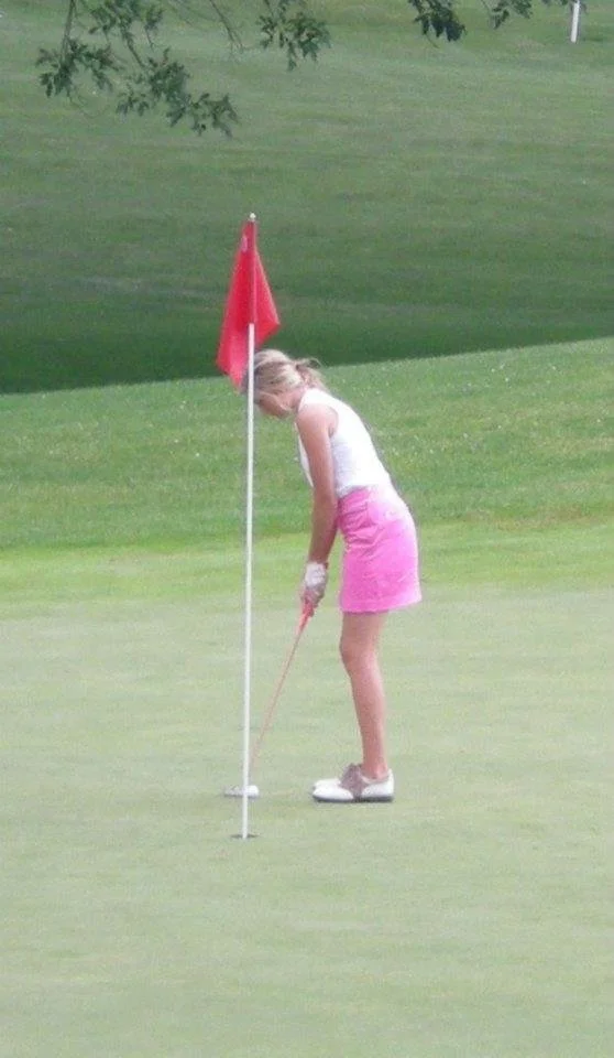 A young Caroline Shinkle dressed in a white top and pink skirt on a golf course, preparing to putt.