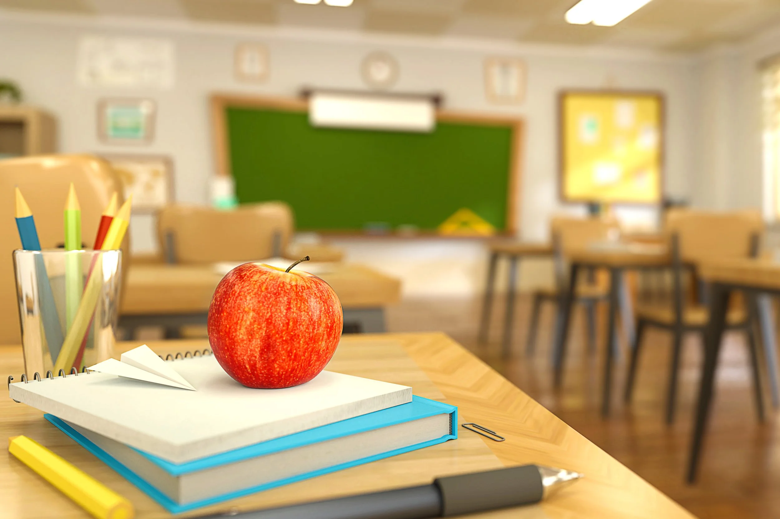 A classroom with a red apple, a notepad, a paper airplane, and a cup of colored pencils on a wooden desk in the foreground. The background features a green chalkboard, chairs, and windows in a softly lit classroom.