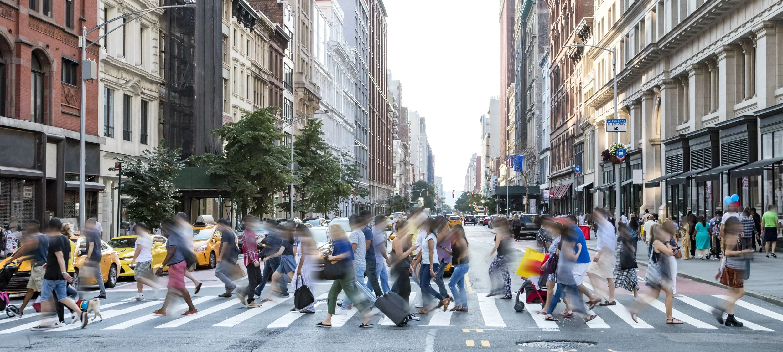 A busy city street with many pedestrians crossing the crosswalk, some with luggage or strollers. Tall buildings line the street, and there are cars and taxis on the road.