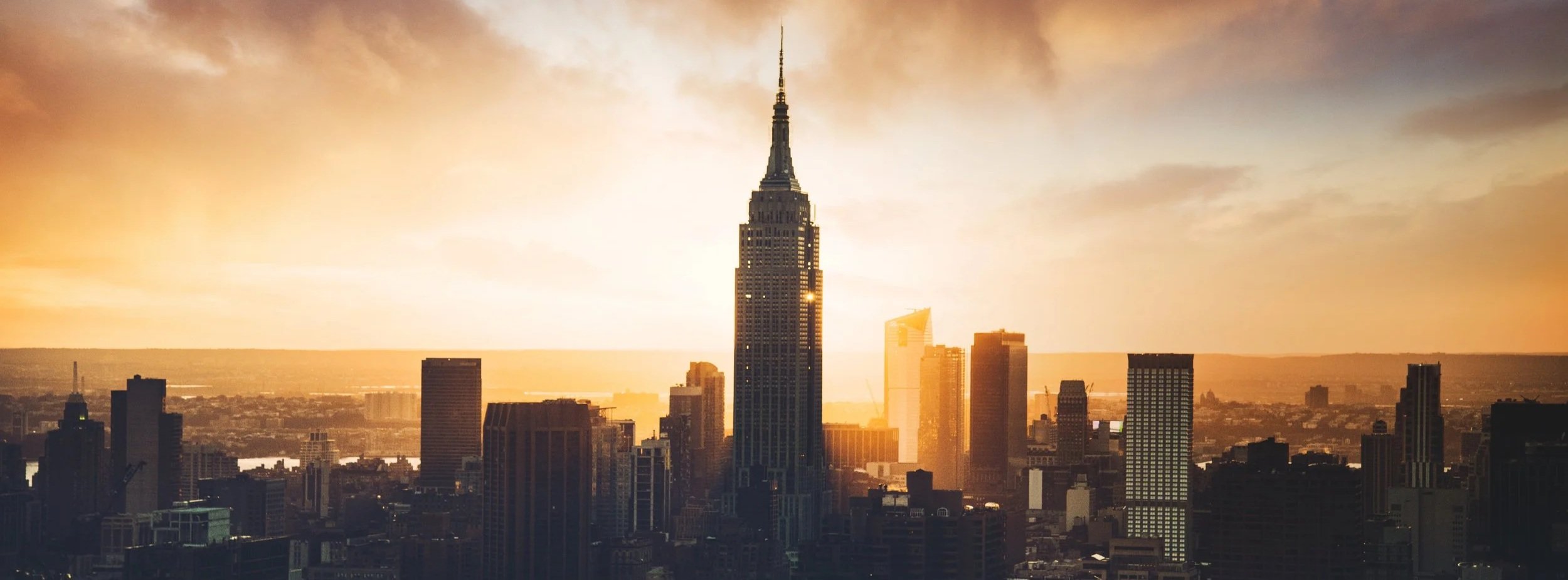 Sunset skyline of New York City featuring the Empire State Building in the center, with the cityscape and water in the background.