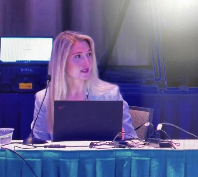 Caroline Shinkle sitting at a desk with a laptop, wearing a blazer, and speaking into a microphone.