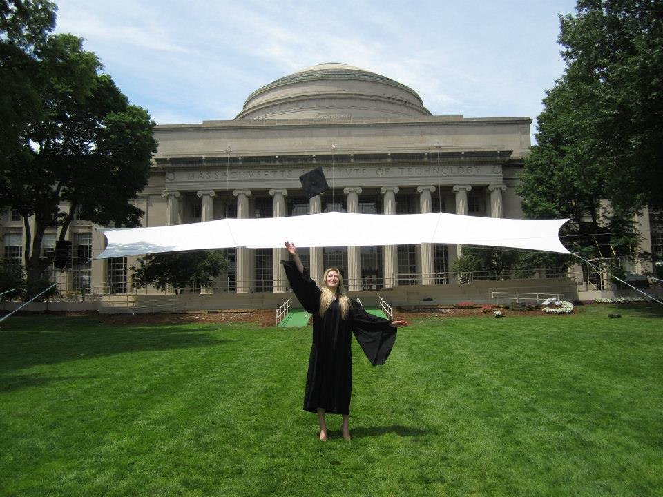Caroline Shinkle in a graduation gown and cap standing on grass in front of a large academic building, throwing her cap into the air with a joyful expression.
