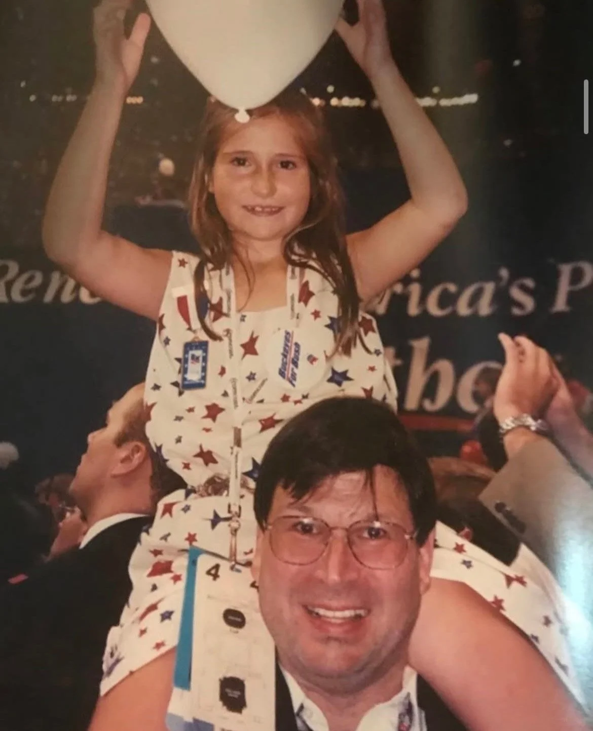 Caroline Shinkle riding on the shoulders of a her father, both wearing matching patriotic-themed outfits with red, white, and blue stars. The girl is holding a white balloon above her head and smiling.