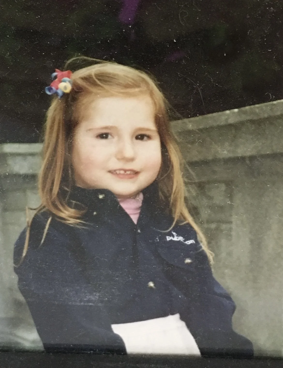 A young Caroline Shinkle with red hair and a colorful hair clip, smiling at the camera, wearing a dark jacket and standing outdoors near a concrete wall.