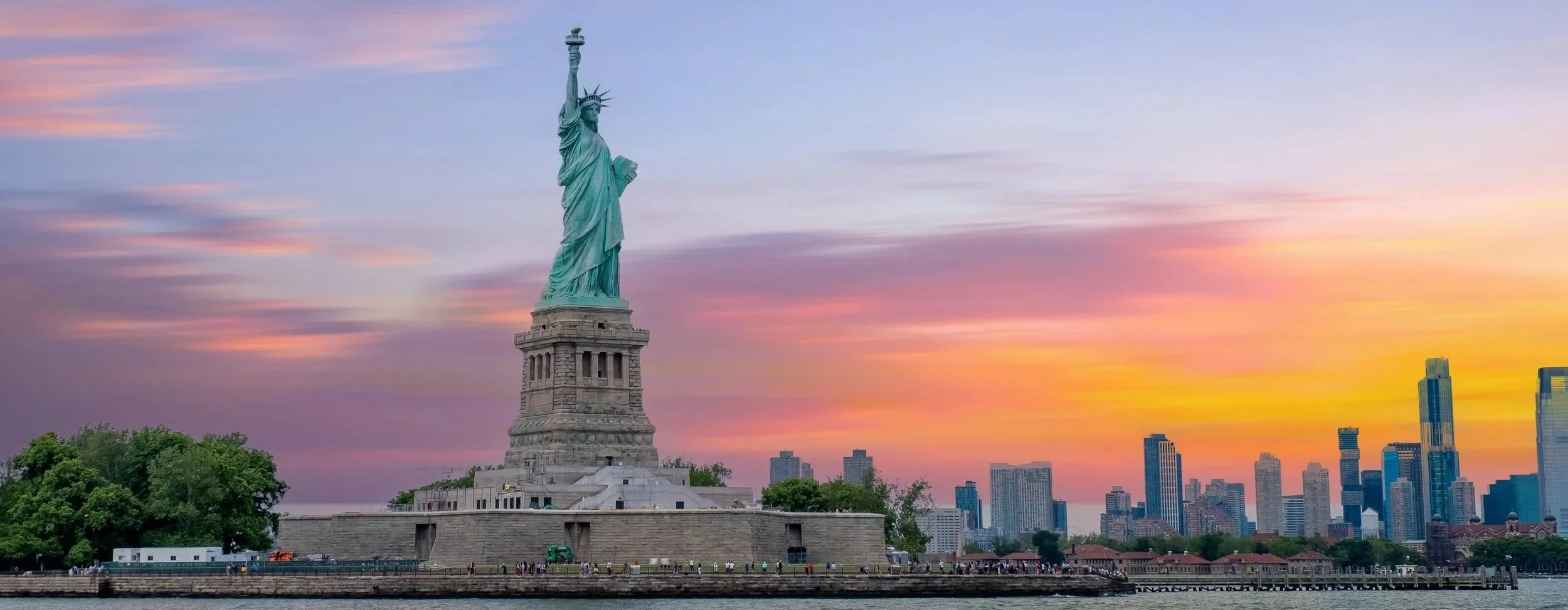 A view of the Statue of Liberty at sunset with a city skyline in the background.