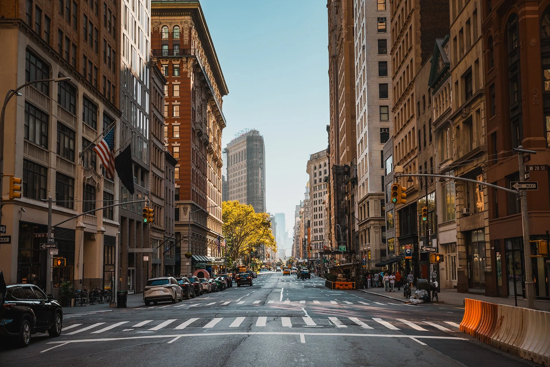 Urban street scene in New York City with tall buildings, cars parked along the street, and a clear sky.
