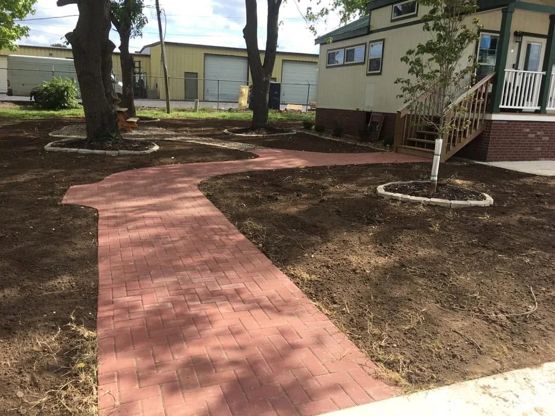 Brick sidewalk winding through freshly tilled soil in front of a house with trees and a yard.