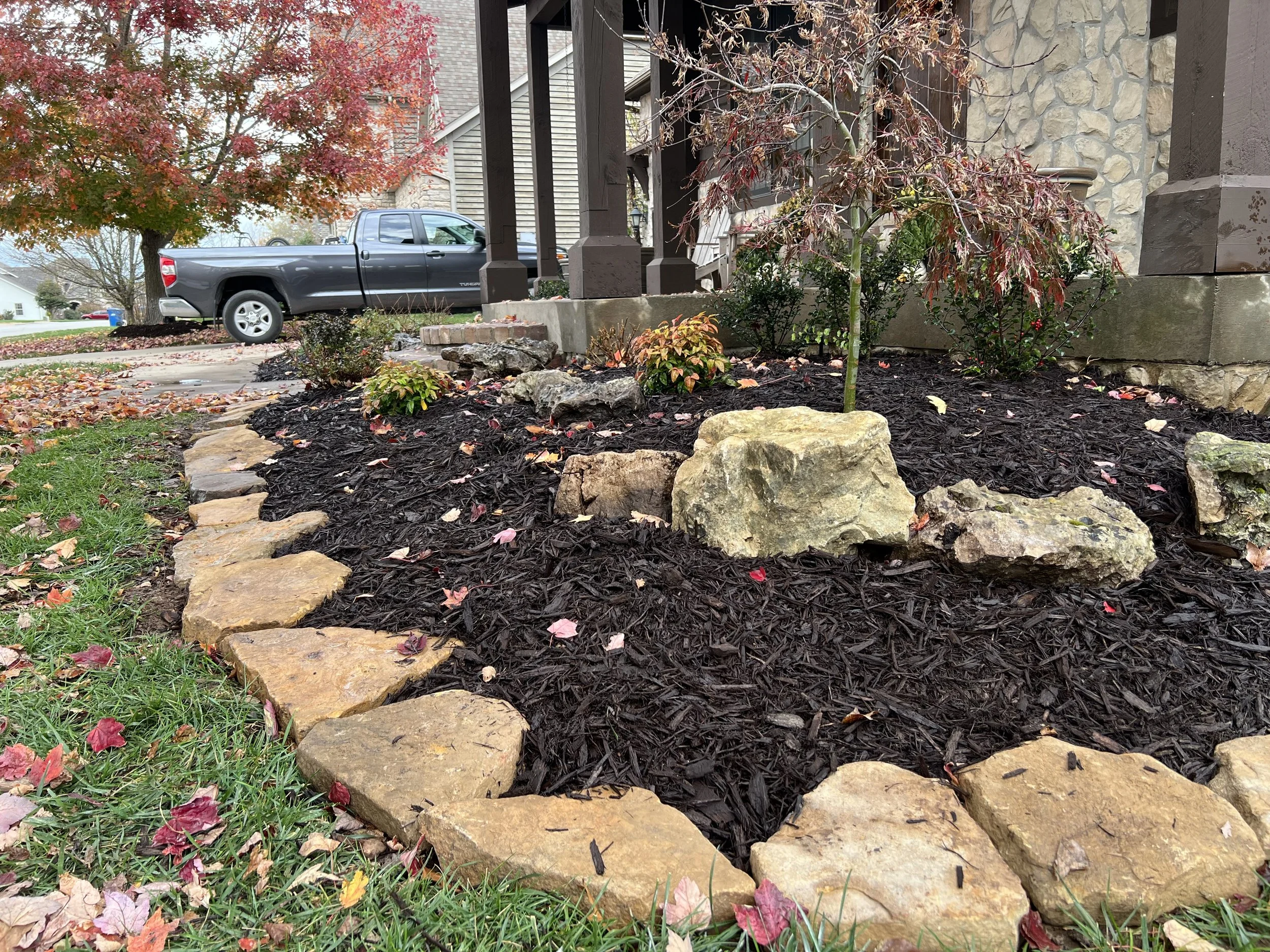 A landscaped front yard with a flower bed bordered by large stones, containing mulch, small plants, and a small tree, near a house with stone and wooden siding, with a gray pickup truck parked on the street in the background.
