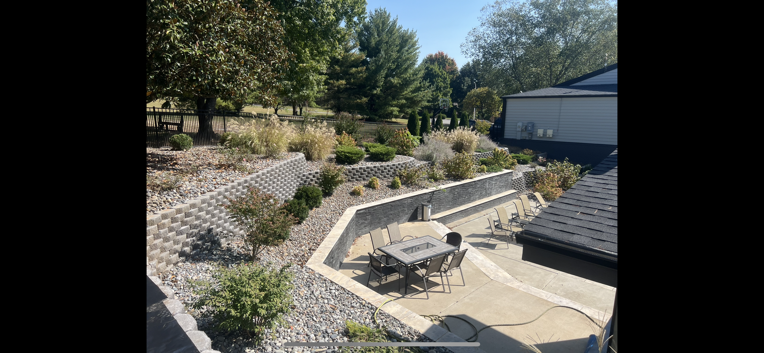 Backyard patio with chairs, table, fire pit, planted landscaping, and a house with a sloped roof, surrounded by trees and a fence.