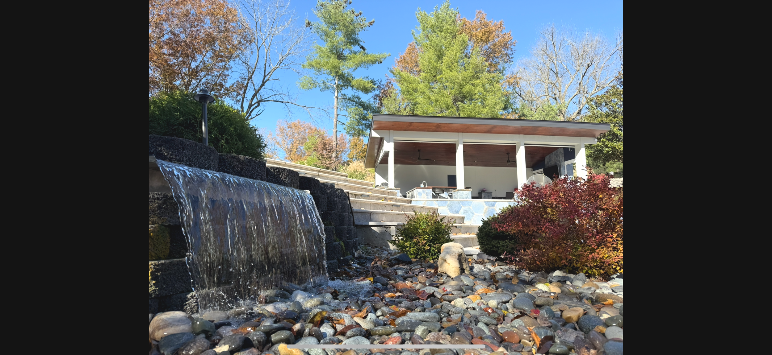 A landscaped backyard featuring a waterfall cascading over dark rocks, surrounded by colorful autumnal bushes and pebbles, with a modern open-air outdoor patio in the background under a clear blue sky.