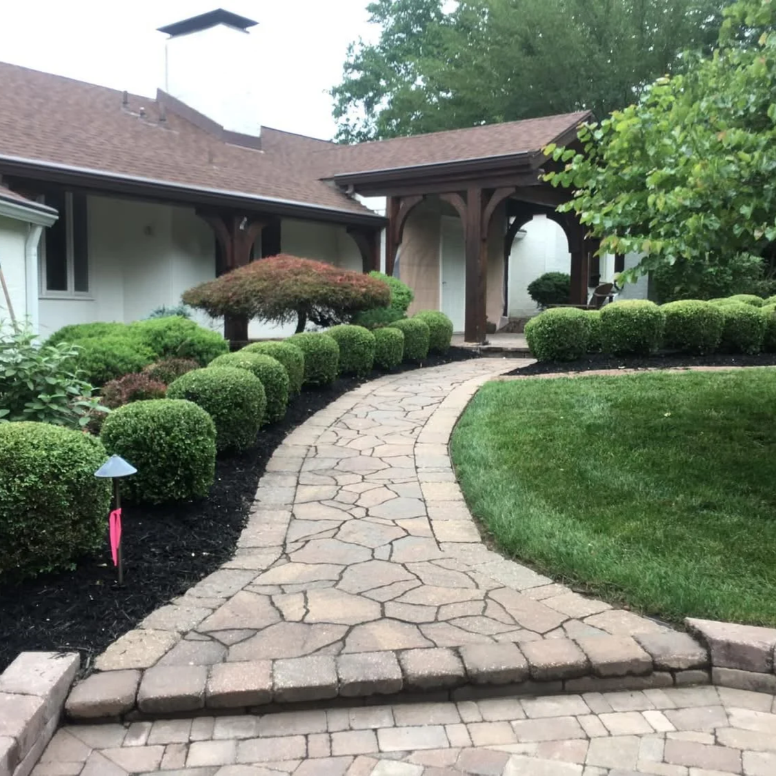 A landscaped front yard with a stone pathway leading to a house, surrounded by neatly trimmed bushes and a green lawn.