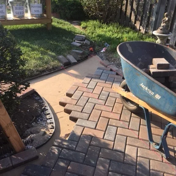 A small outdoor patio area with a partially completed brick walkway, a blue wheelbarrow filled with bricks, and garden supplies in the background.