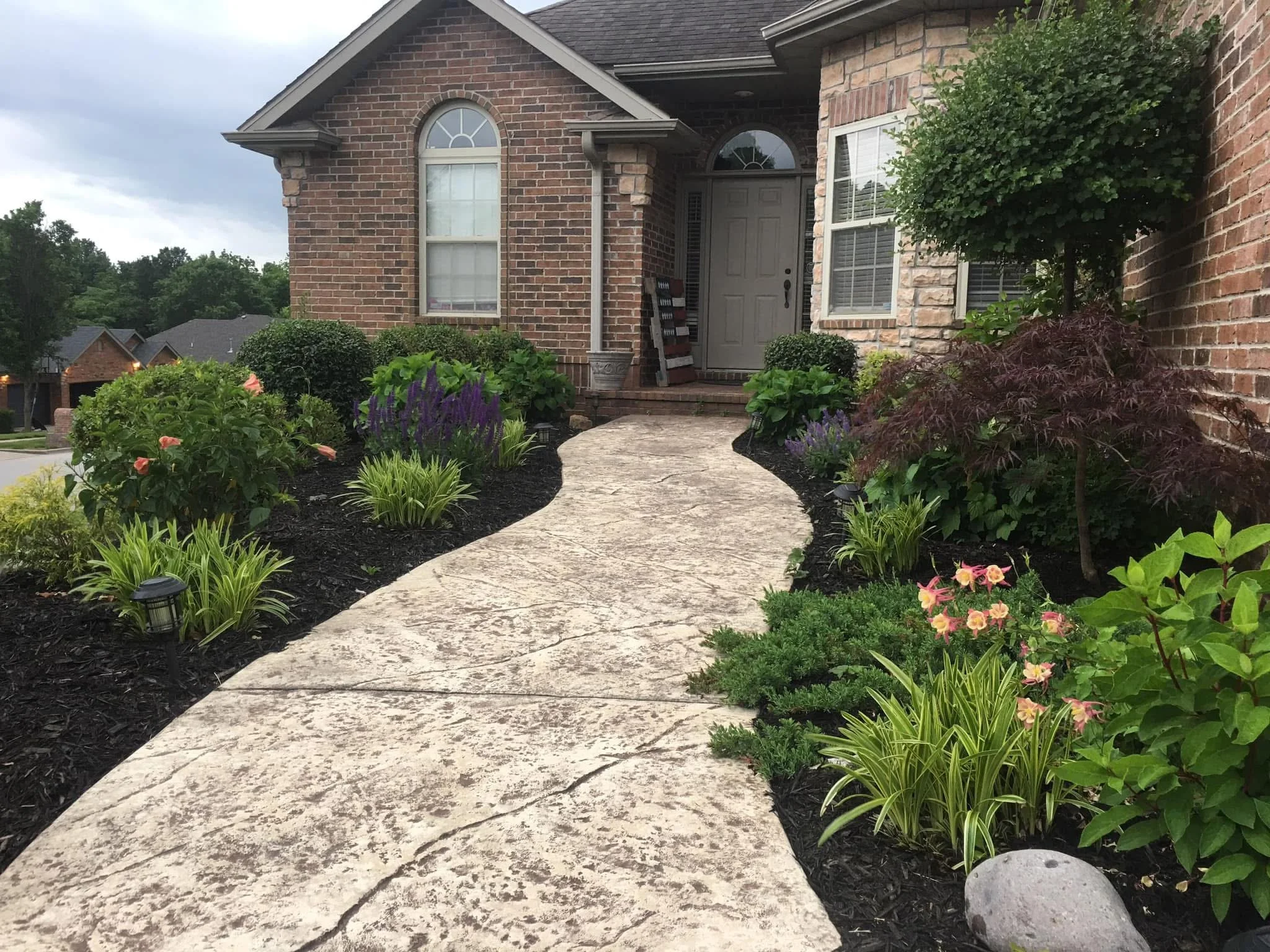 A stone pathway leading to a front door of a brick house, surrounded by well-maintained flower beds with colorful plants and shrubs.