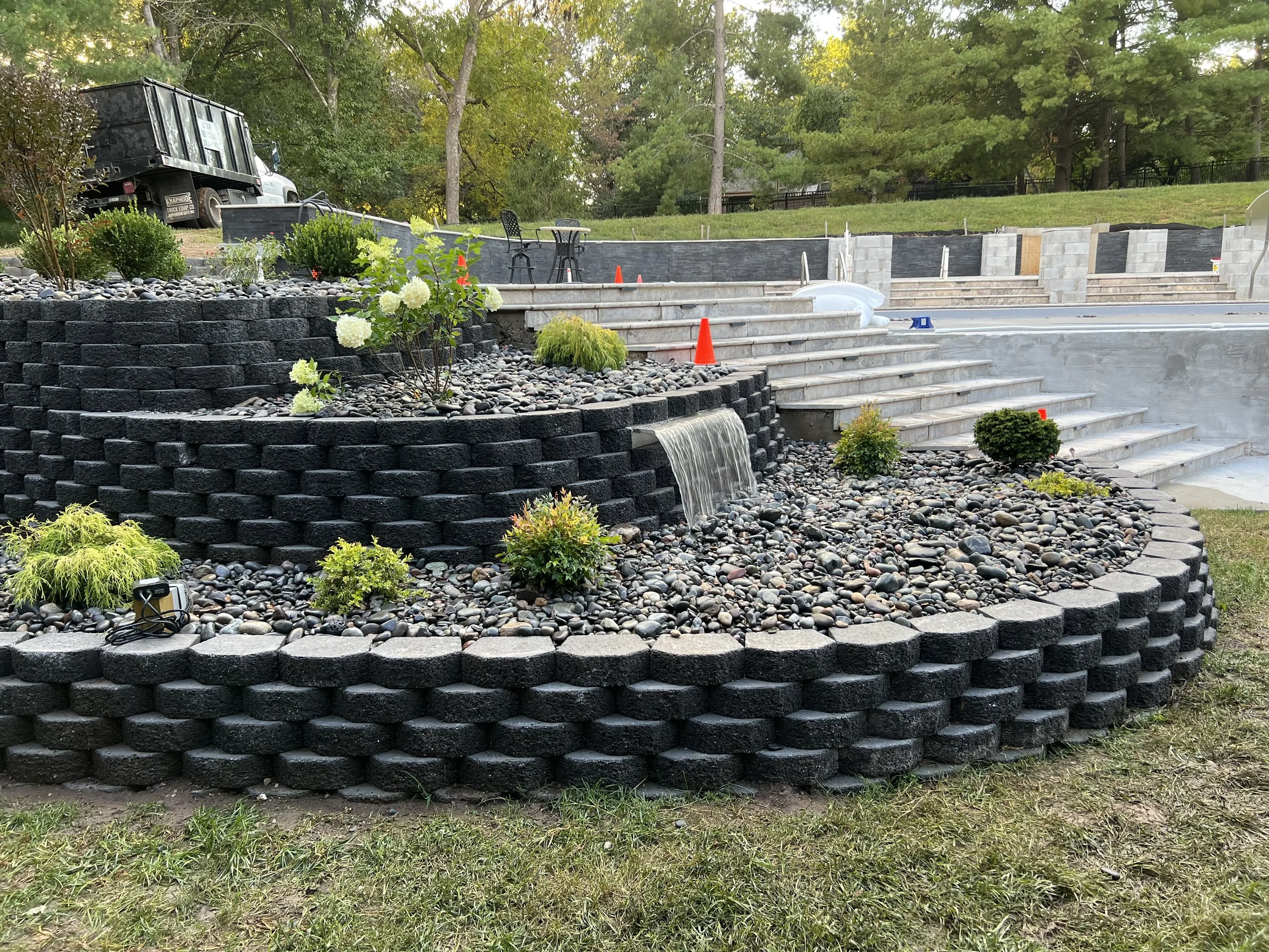 Landscape of an outdoor construction site featuring layered black brick retaining wall with small plants and a small waterfall, construction cones, and concrete stairs under development, surrounded by trees.