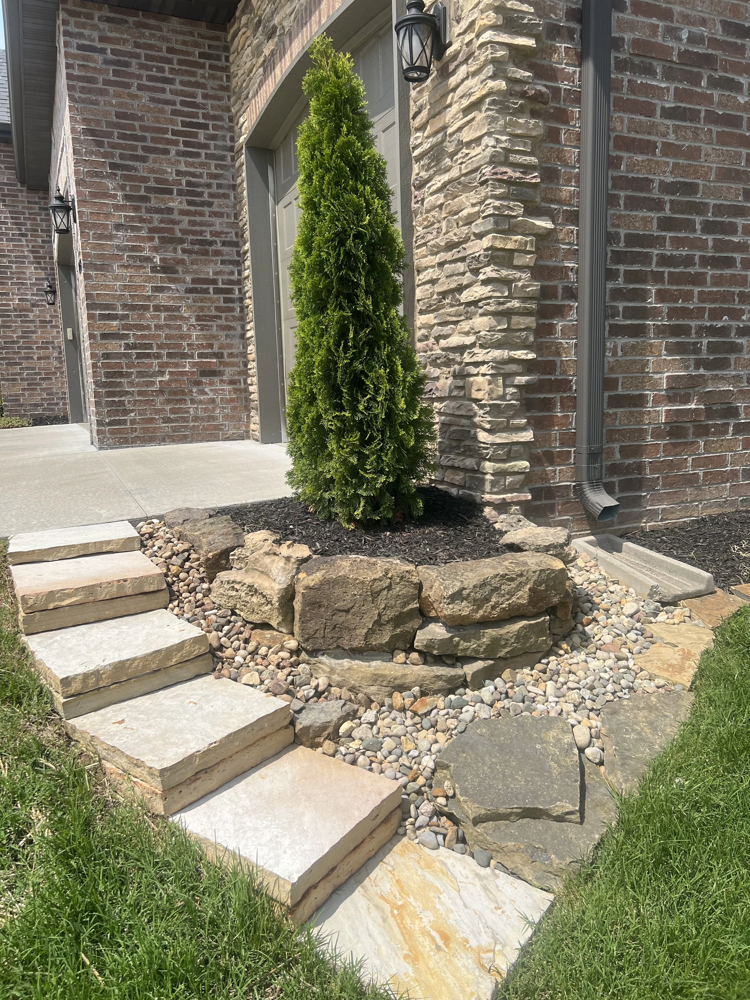 A small, neatly landscaped front yard featuring a tall, narrow green shrub, surrounded by large rocks and smaller decorative stones, with a stone pathway leading to a house with a brick exterior.