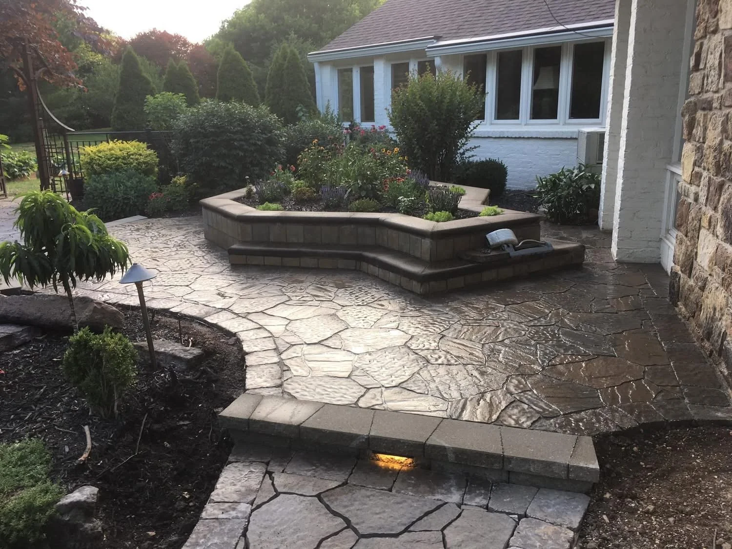 Backyard patio with a stone pathway, raised garden bed with plants and flowers, surrounding greenery, and a house wall with windows.
