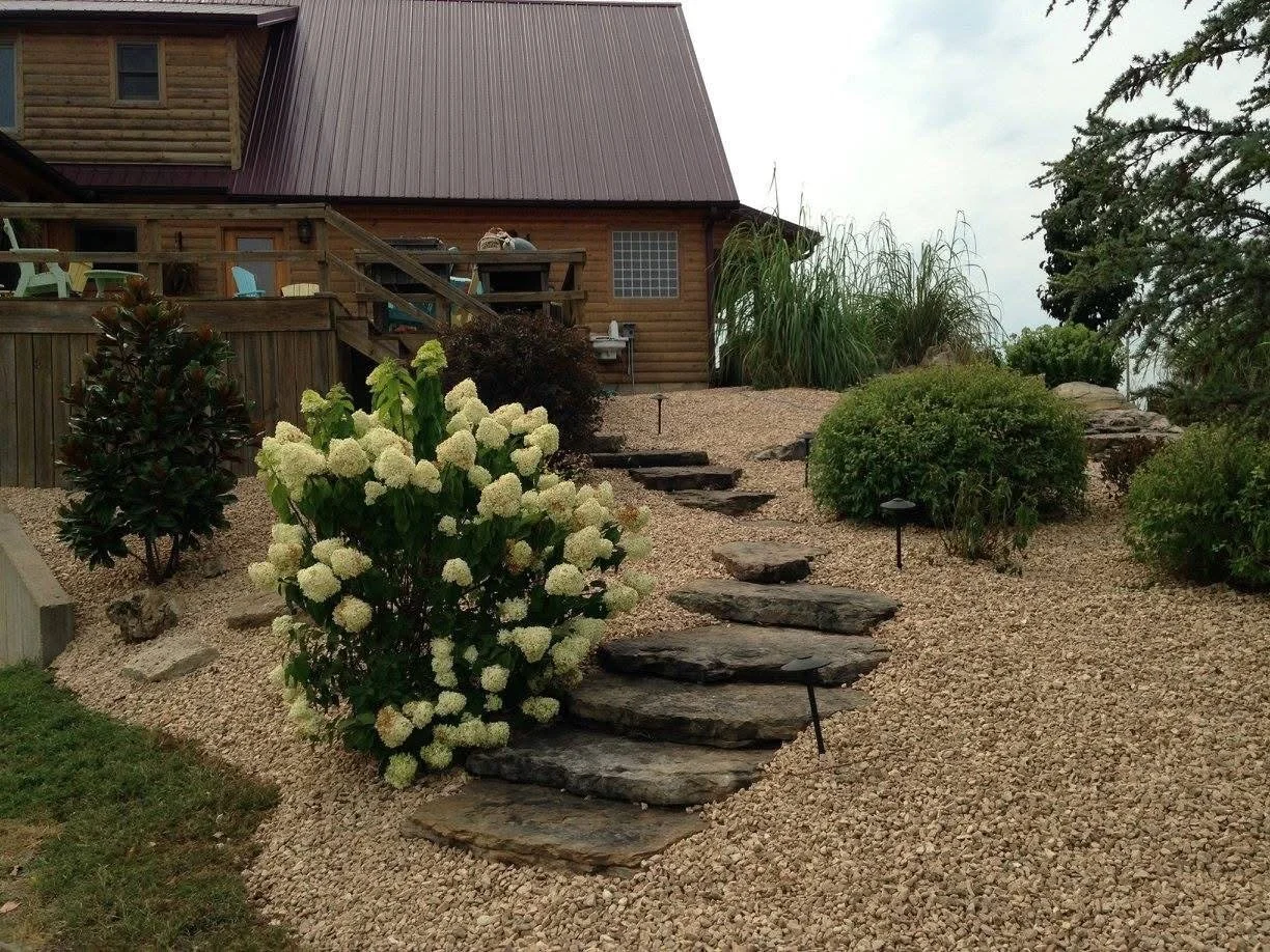 A gravel landscaped yard with a stone pathway leading to a wooden house with a deck, surrounded by various bushes and plants, on an overcast day.