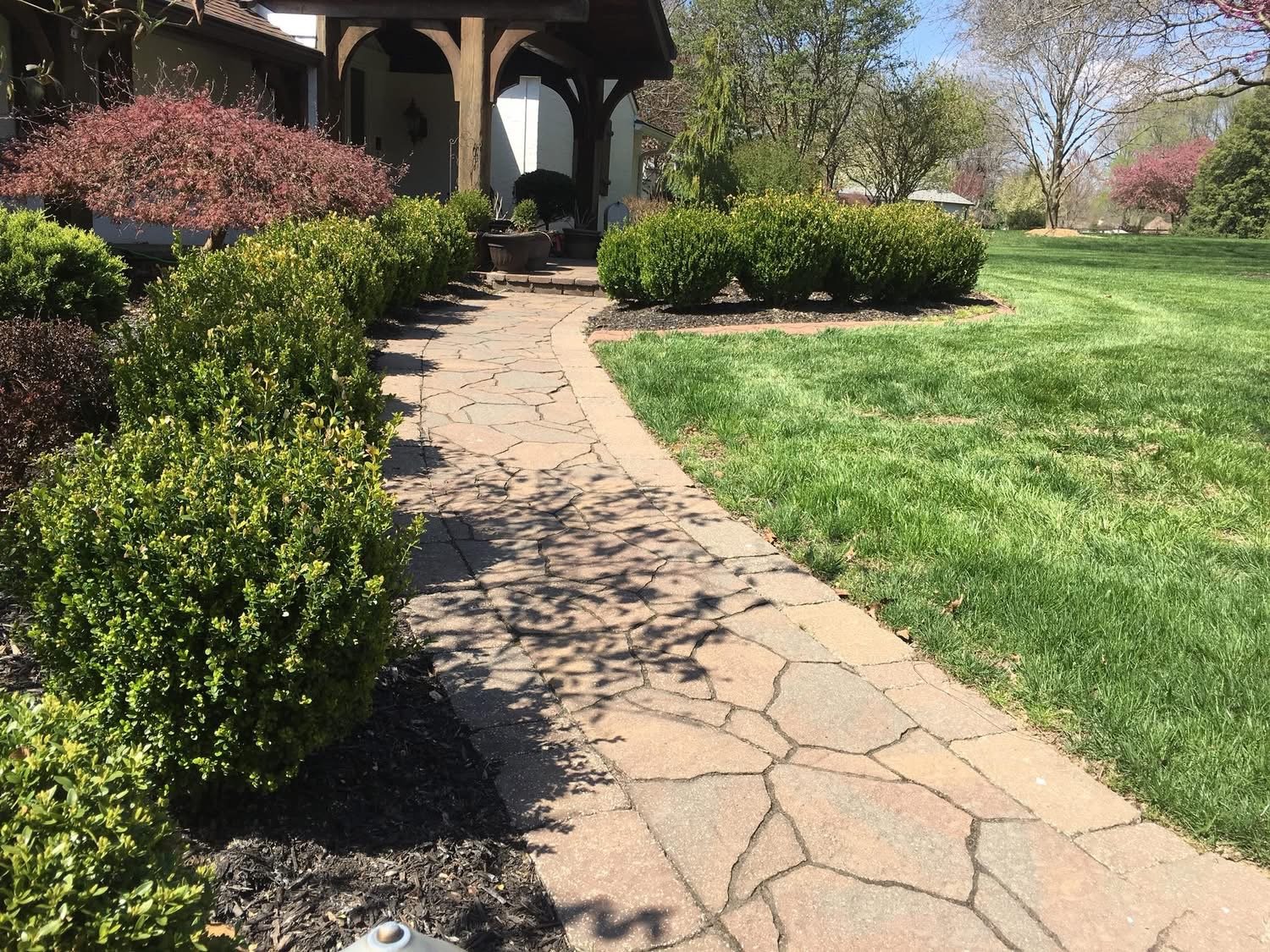 A stone pathway leading to a porch of a house, surrounded by green bushes and a grassy lawn with trees in the background.