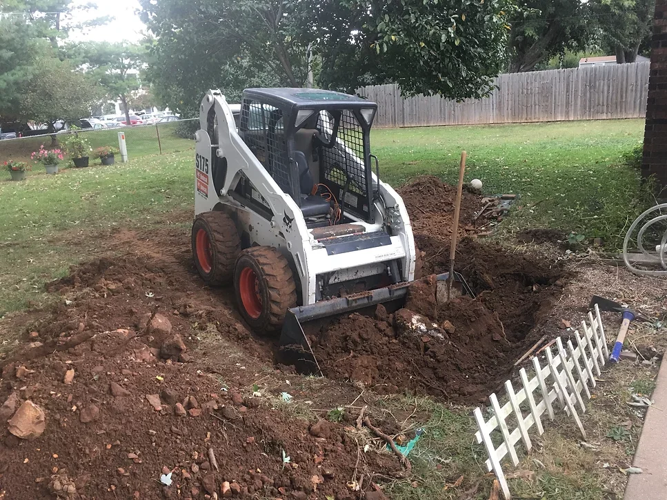Mini skid steer loader digging a hole in a backyard with a small white picket fence nearby.
