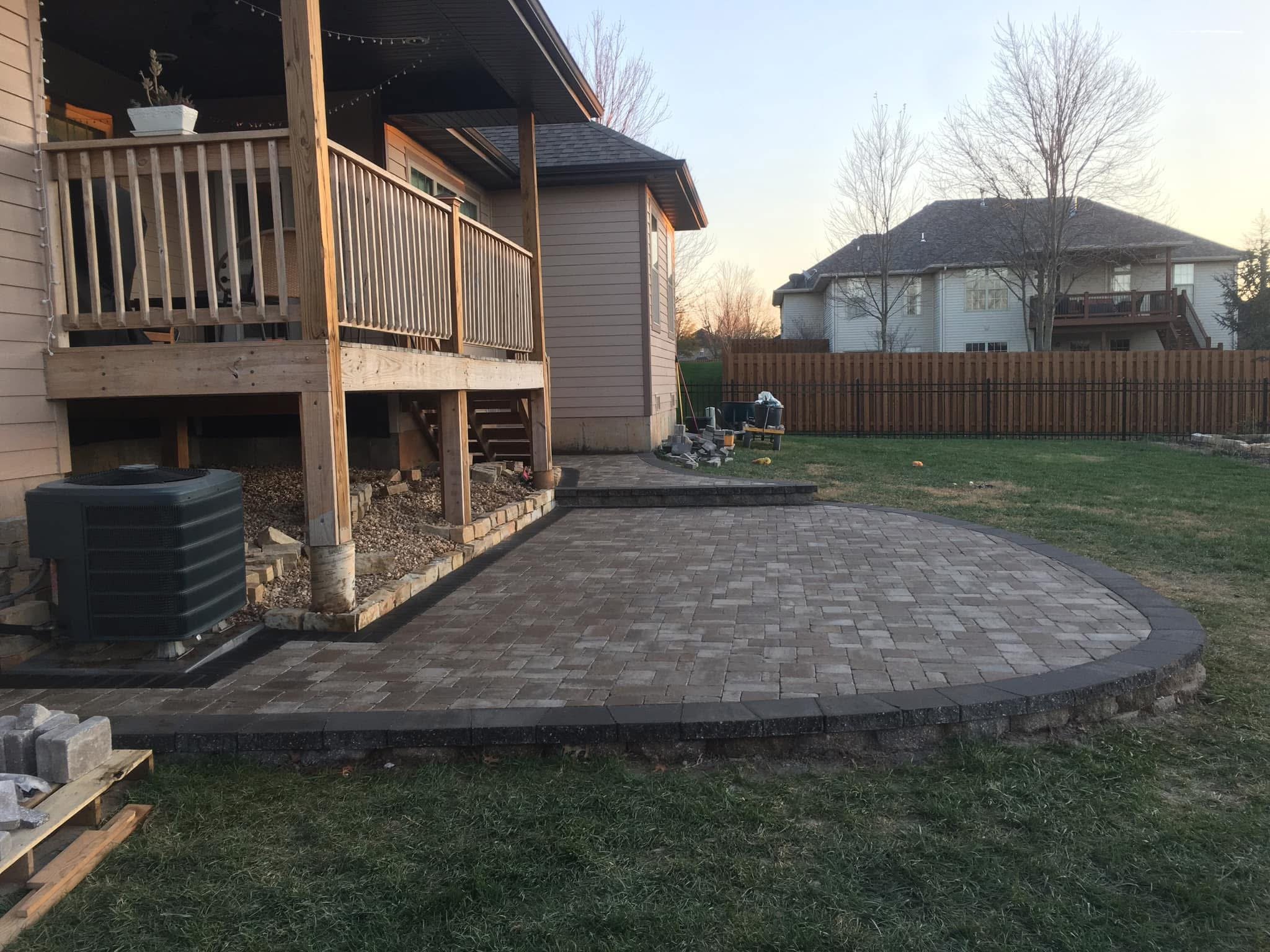 Backyard with newly constructed brick patio, a wooden deck with stairs, and a fenced yard with neighboring house and trees.
