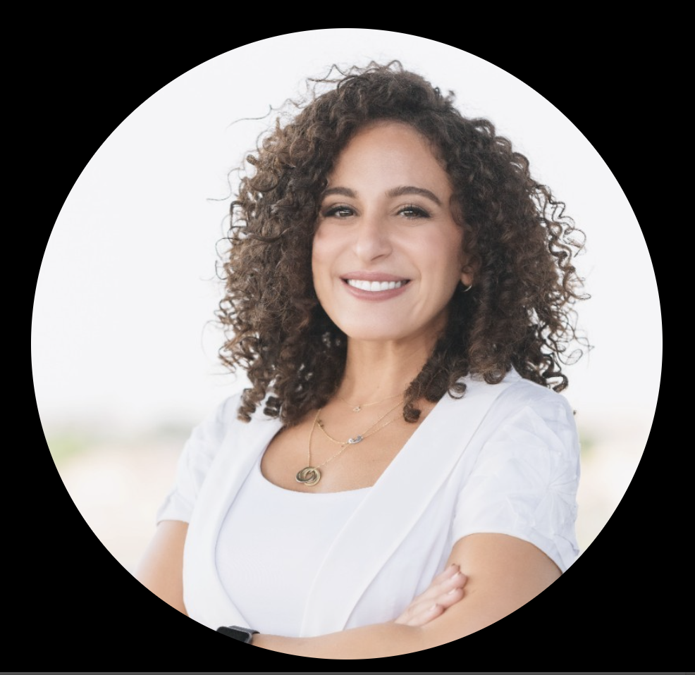 A woman with curly brown hair smiling, wearing a white top and jewelry, crossing her arms. Background is blurred with a light sky.