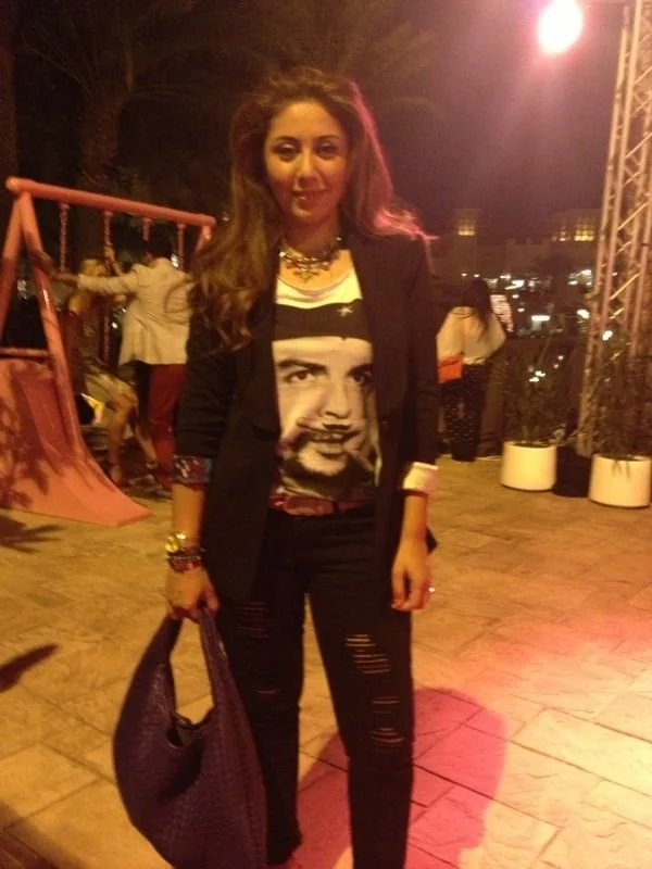 A woman with long, voluminous hair standing outdoors at night with warm lighting, wearing a graphic t-shirt with a portrait, a black blazer, distressed black jeans, and holding a large bag.