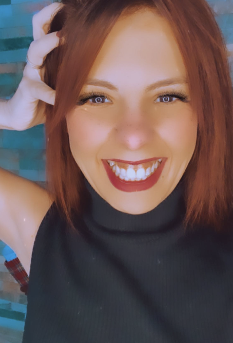 Close-up of a smiling woman with red hair, wearing a sleeveless black top, in front of a brick wall.