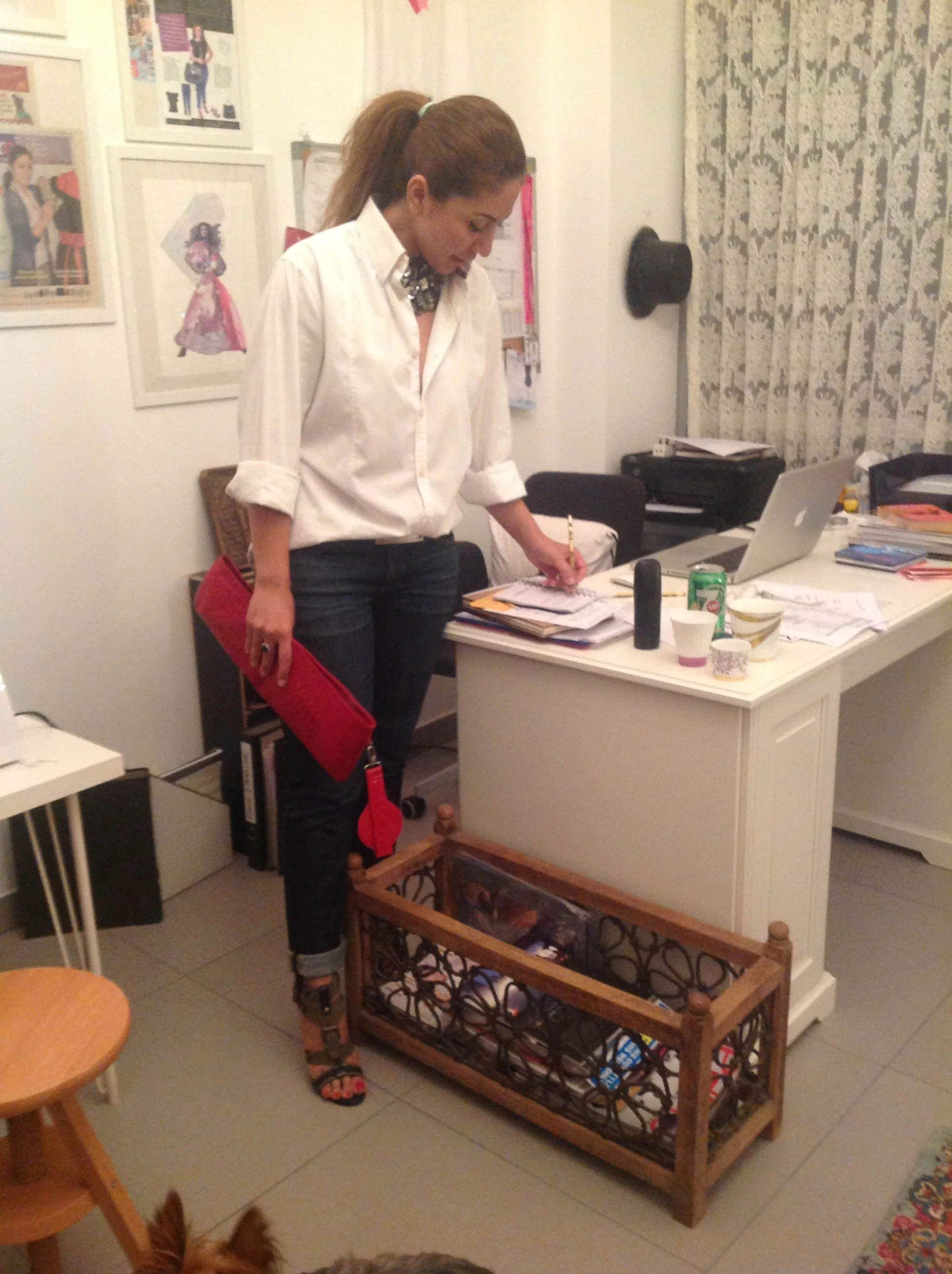 A woman dressed in a white shirt and dark jeans is standing in an office, leaning over a cluttered desk while holding a red clutch purse. The desk has papers, a laptop, and various cups on it. There is a wooden pet crate filled with miscellaneous items on the floor in front of her, and a small dog is partially visible at the bottom edge of the image.