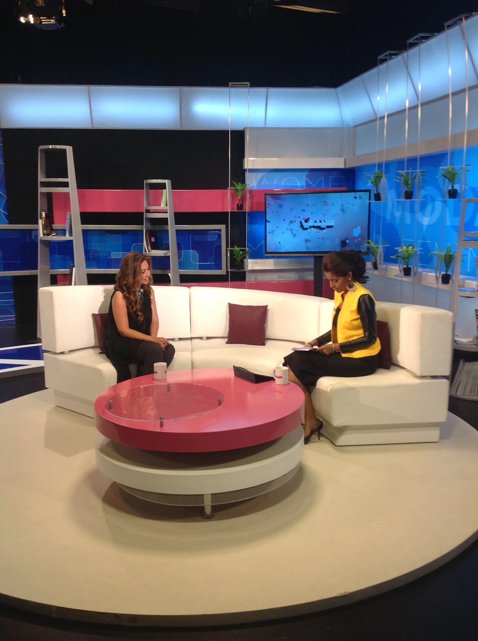 Television studio set with two women seated on a white sofa, a woman in black and another in yellow. The set includes a pink circular table, decorative plants, and a large screen in the background.