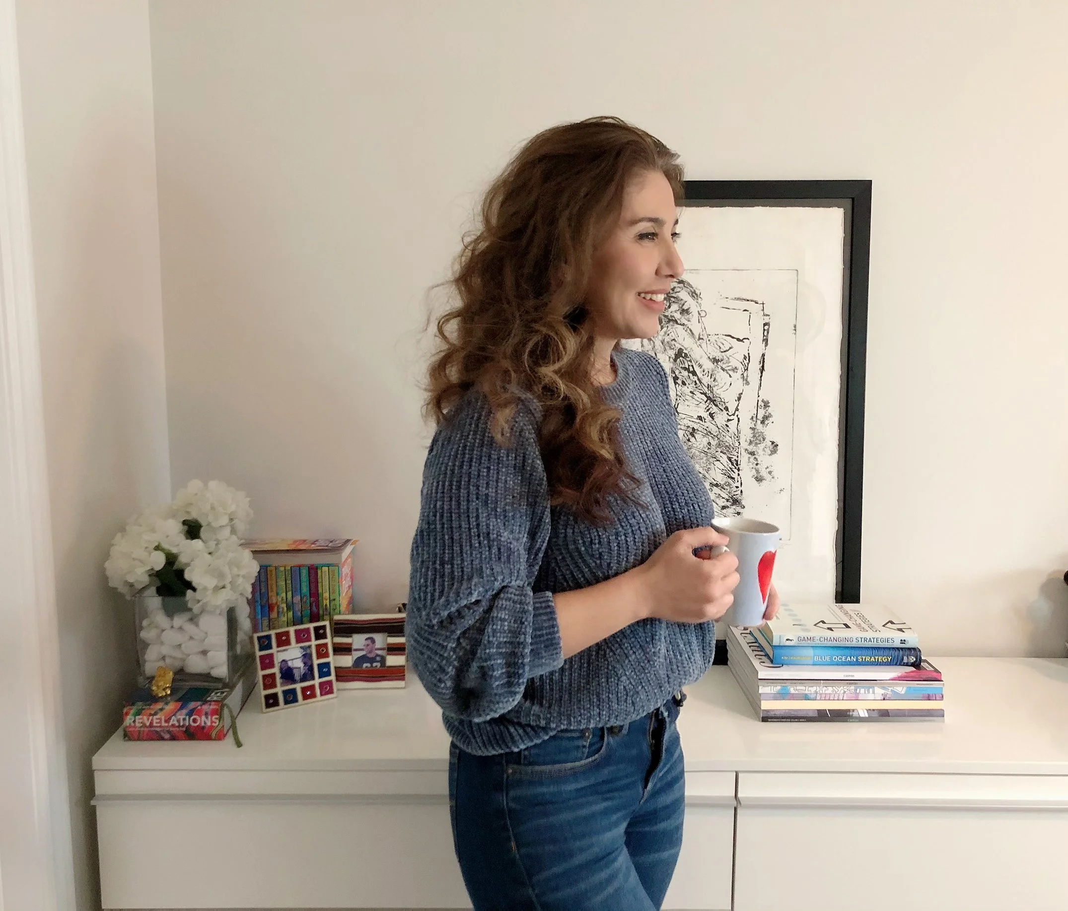A woman with curly red hair holding a white mug with a red logo, standing in a room with a white sideboard and various books and framed photographs on top, smiling and looking to her right.