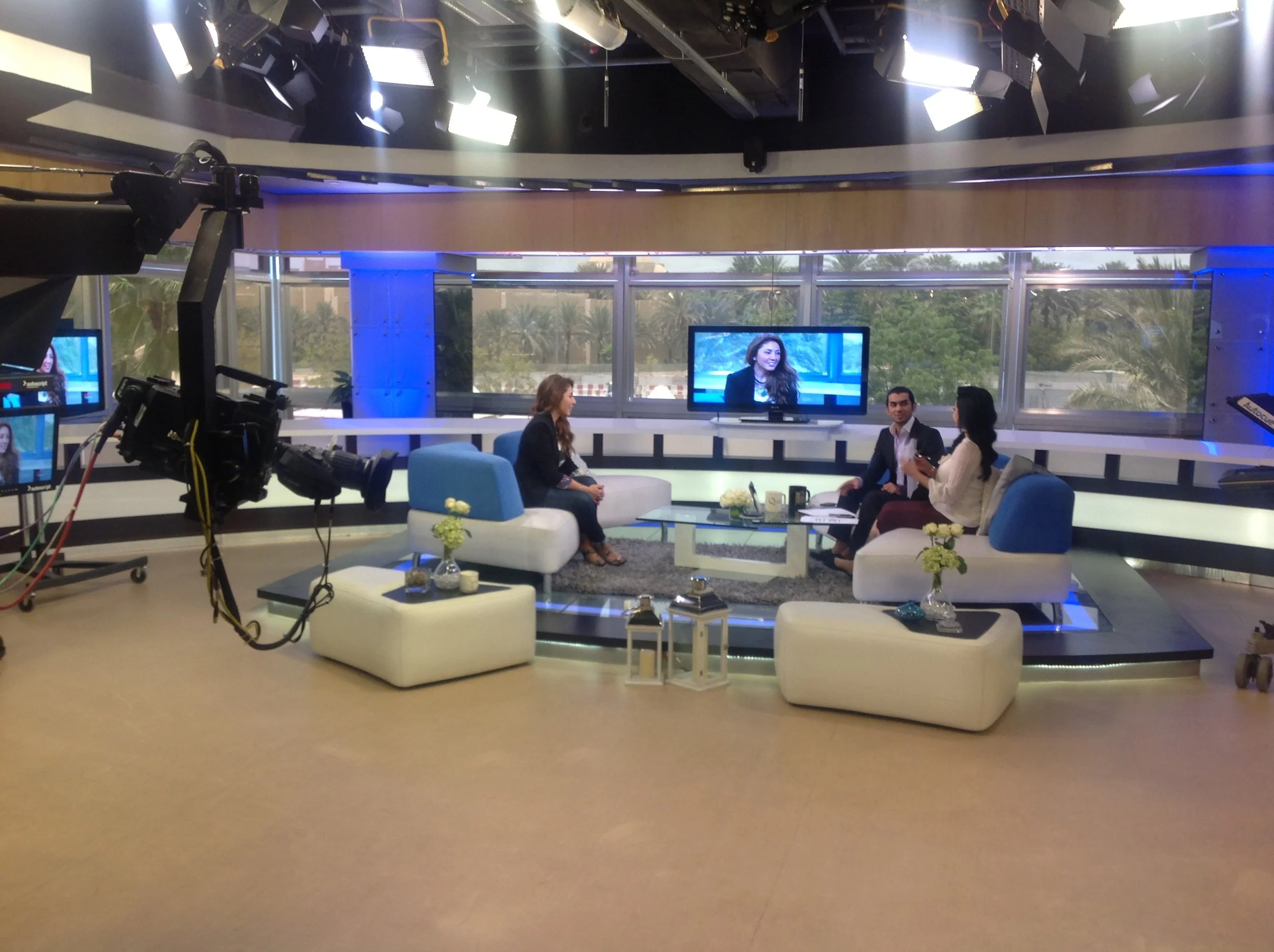 TV studio set with two hosts interviewing Rana AlBasri Mouawad on sofas and chairs, studio lights, a camera on the left, and large windows showing a sunny outdoor view with palm trees.