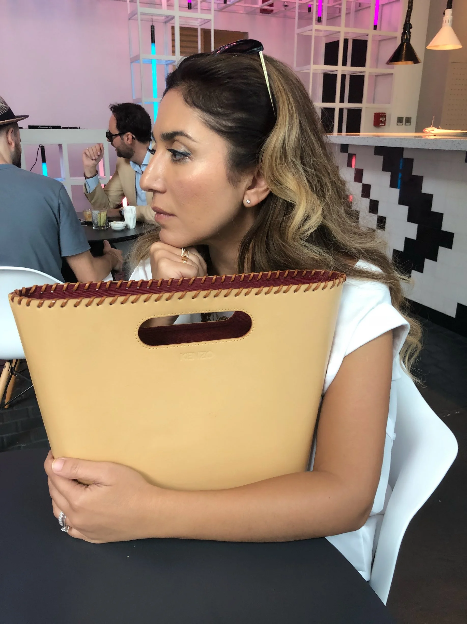 A woman with wavy hair and sunglasses on her head sitting at a table in a modern restaurant, holding a large yellow bag, with a thoughtful expression.