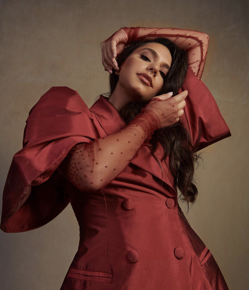 A woman with long dark hair wearing a red satin dress with sheer polka-dot sleeves, posing with her hand on her head and arm raised, against a plain background.