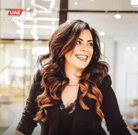 A smiling woman with long wavy brown hair and a black blazer, sitting in a modern office environment.