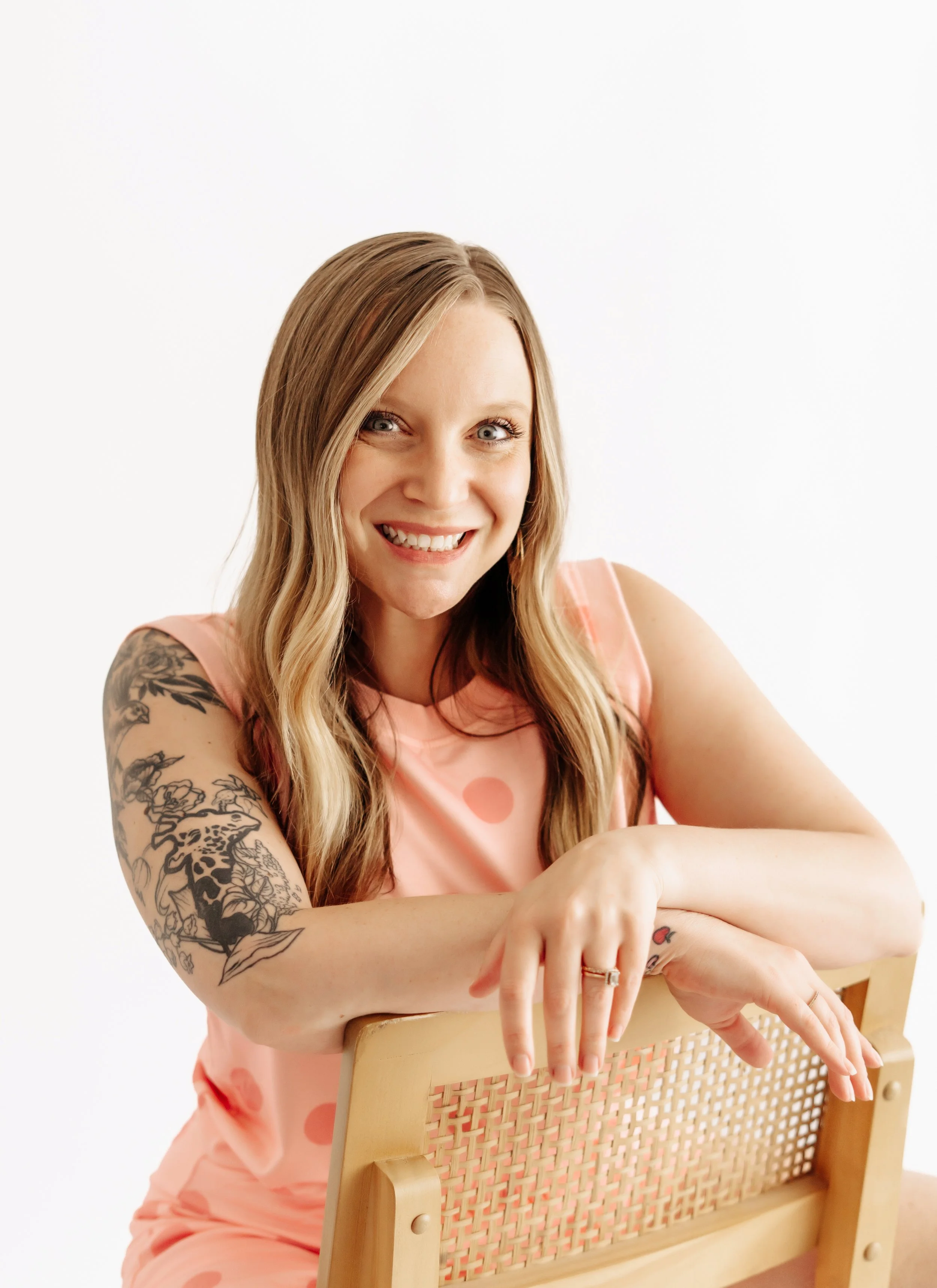A smiling woman with blonde hair and tattoos, wearing a pink polka dot shirt, sitting behind a wooden chair with a woven backrest, pointing at her wrist."}