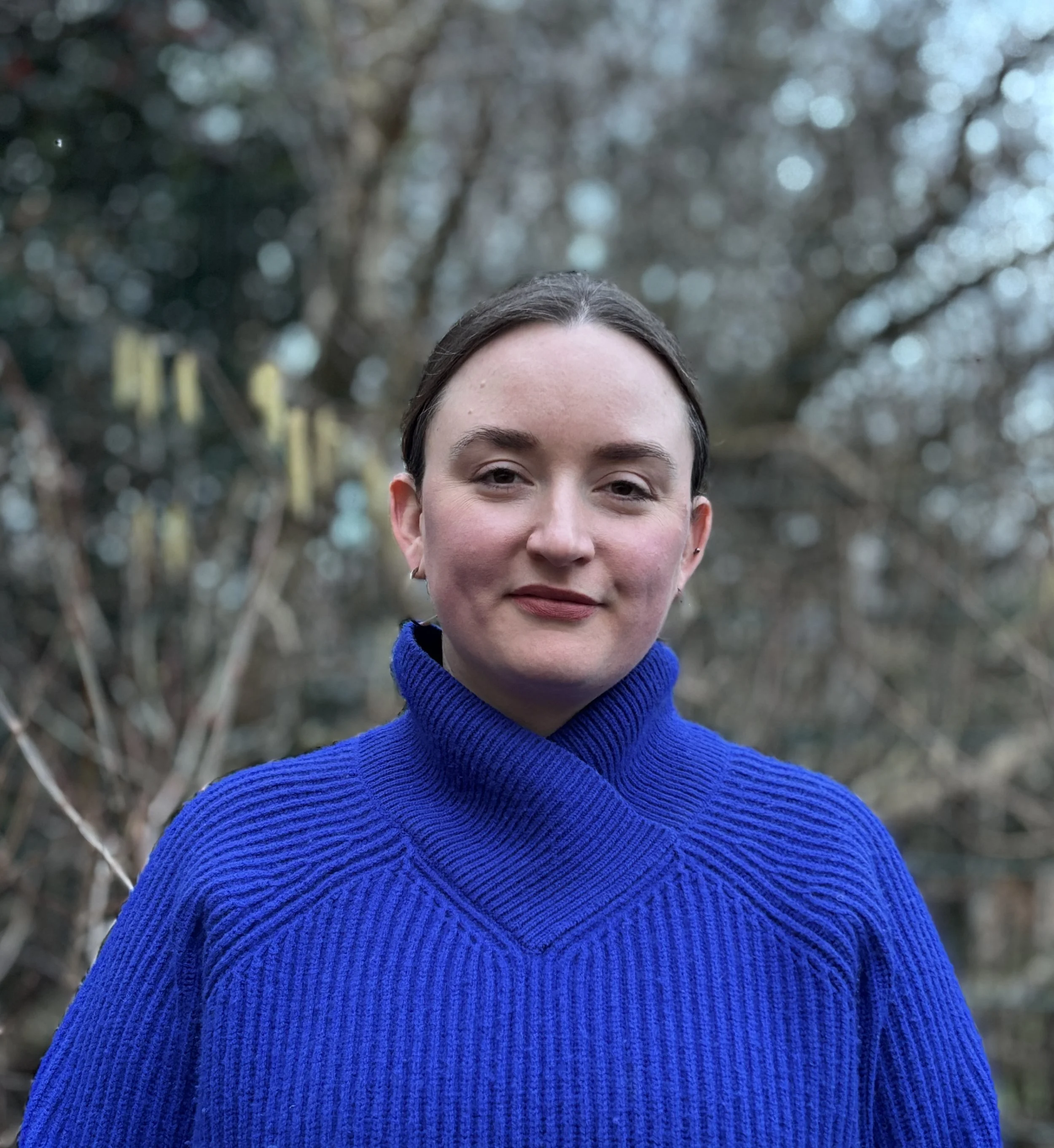 A woman with dark hair in a bun, wearing a bright blue turtleneck sweater, standing outdoors in front of a blurred background of trees and branches.