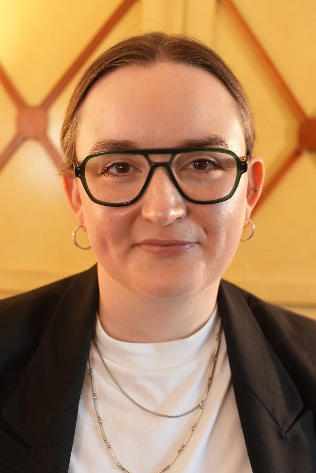 Close-up of a woman with brown hair, glasses, hoop earrings, smiling, wearing a white shirt layered with jewelry, and a black blazer, background with a wooden ceiling.