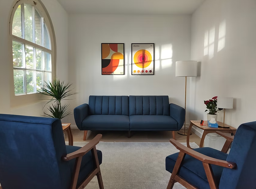 Living room with a navy sofa, two matching navy armchairs, a wooden side table with a potted plant, and a white floor lamp. Art prints with geometric and colorful designs hang on the white wall. A large window with green foliage outside provides natural light.