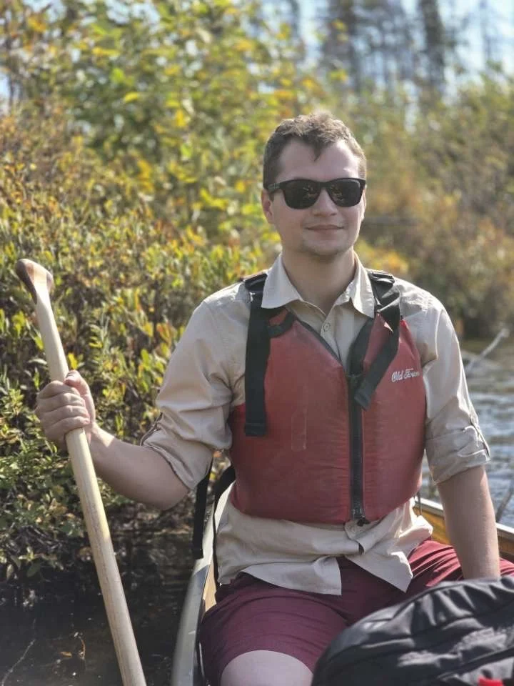 A man wearing sunglasses, a beige shirt, and a red life vest paddling a kayak in a river with autumn-colored trees in the background.