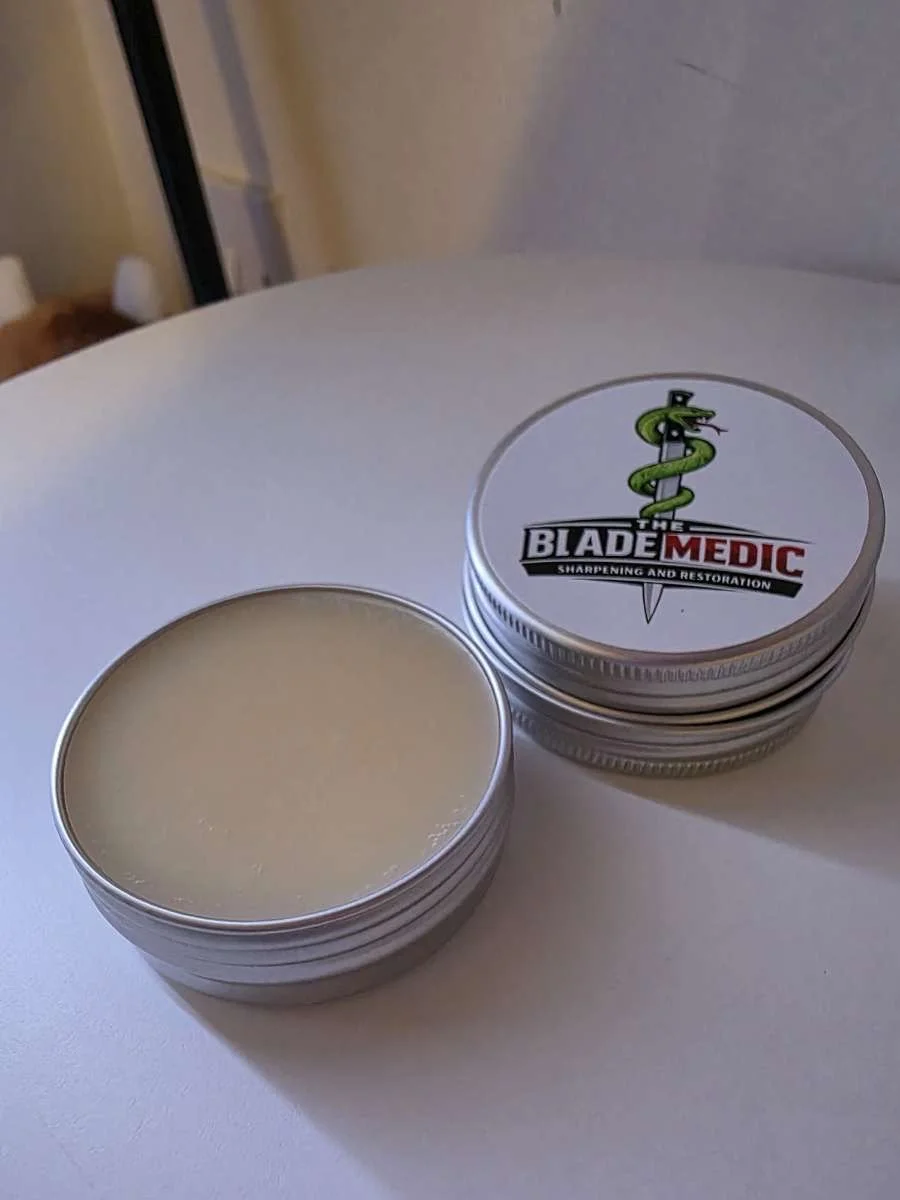 A small round tin container with a lid featuring a logo of a snake wrapped around a sword, labeled 'The Blade Medic' for sharpening and restoration, placed on a white surface.