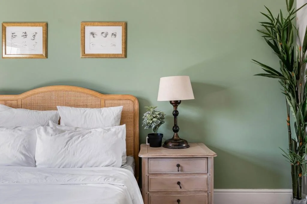 Bedroom with a wooden headboard, white bedding, pillows, a nightstand with a lamp and a potted plant, and framed artwork on a green wall.