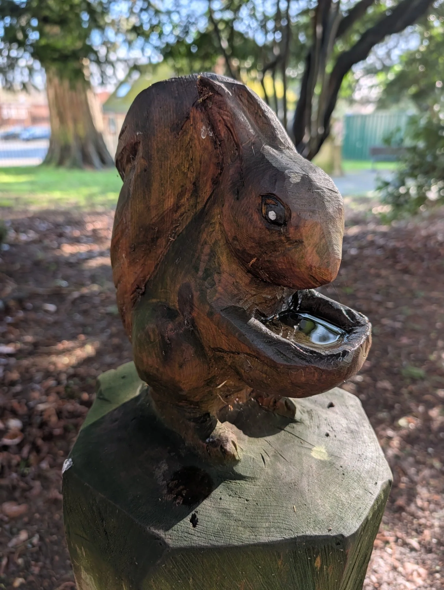 A carved wooden sculpture of an animal, possibly a dog or bear, with an open mouth containing a reflective object, positioned on a wooden pedestal outdoors with trees and greenery in the background.