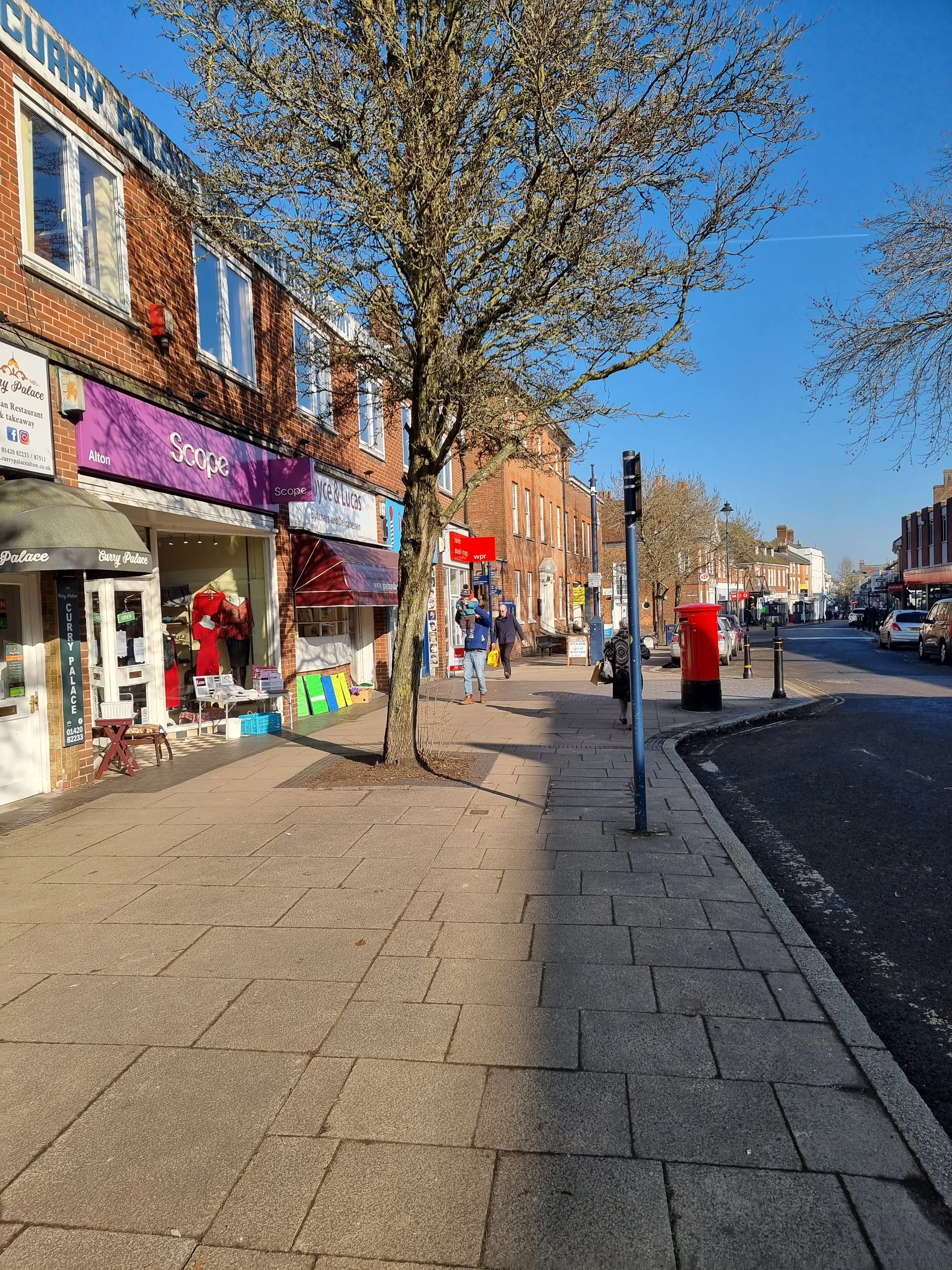 A sidewalk in a small town or city with brick buildings, storefronts, a leafless tree, and pedestrians walking. There is a red post box and a blue parking meter. The sky is clear and sunny.