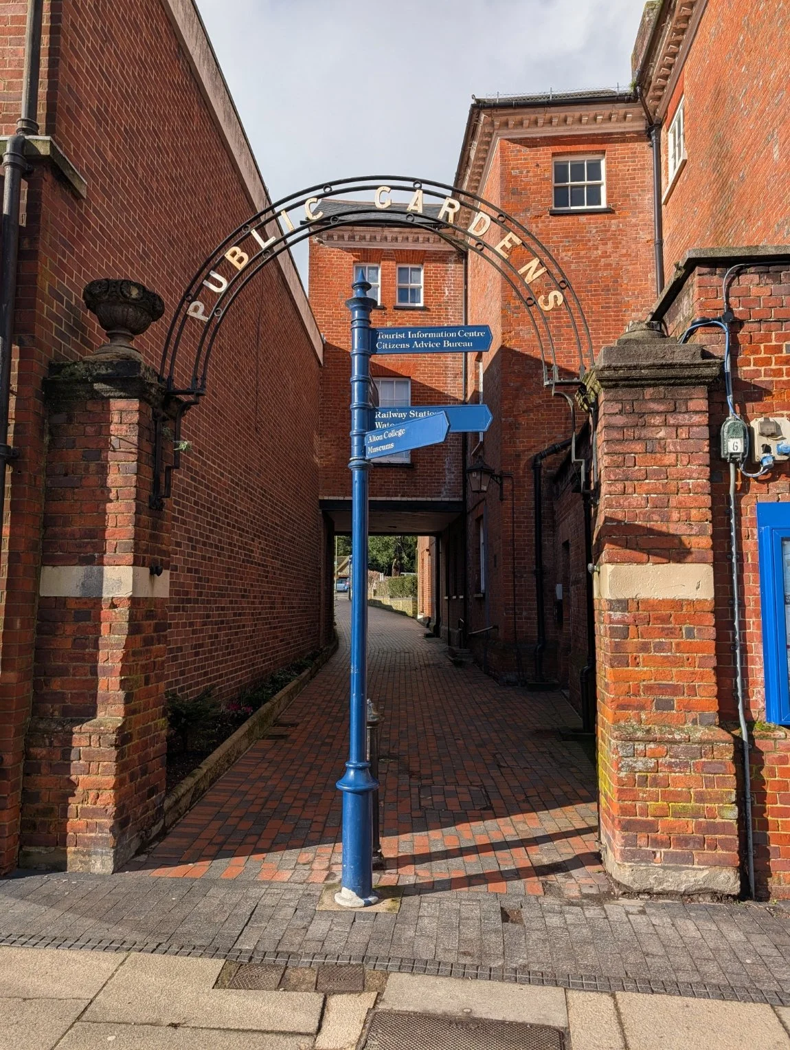 Entrance to Public Gardens with a blue signpost and archway, brick buildings on either side, sidewalk and brick pavement.
