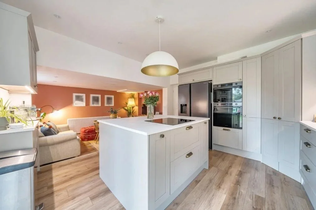 Open concept kitchen with white cabinets, a central island, stainless steel appliances, and a view into the living room with a beige couch, orange accent wall, and decorative plants.