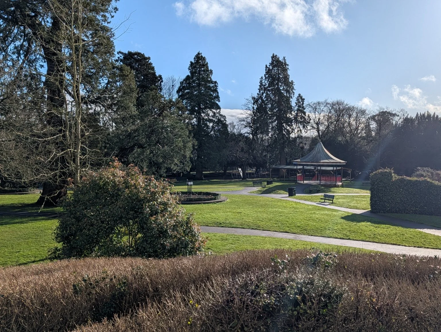 A park with green grass, trees, a gazebo, benches, and clear skies.