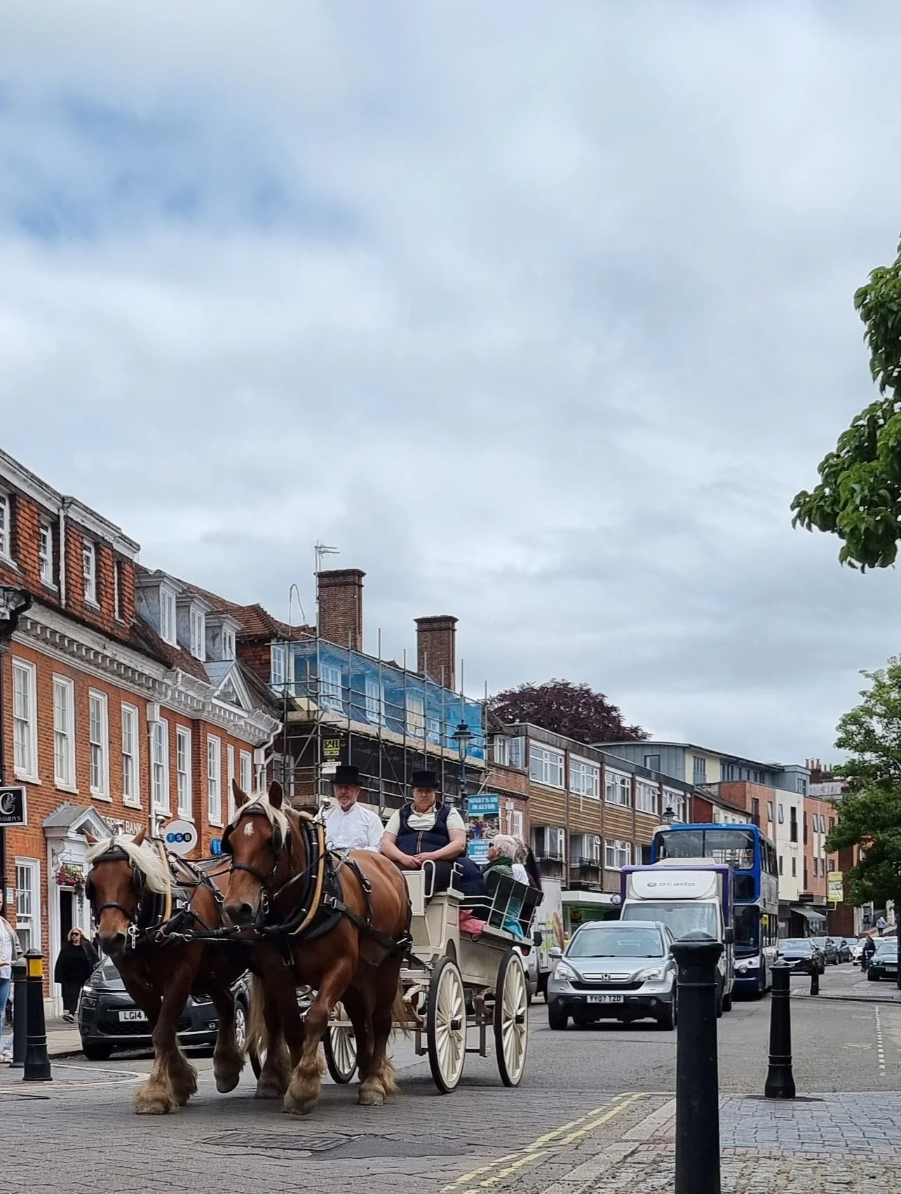 Horse-drawn carriage with two people dressed in vintage clothing, riding through a city street with cars and buildings in the background.